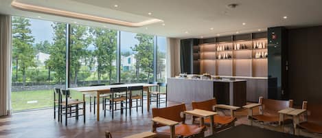 Un comedor moderno con mesas y sillas de madera, una zona de bar con estantes llenos de botellas, y amplias ventanas que ofrecen vistas al exterior.