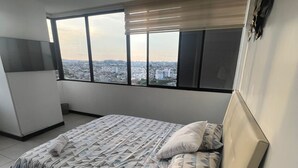 Ein Schlafzimmer mit Bett, einem Fenster mit Jalousien, einer Wanduhr und einem kleinen Tisch mit Fernbedienung.
