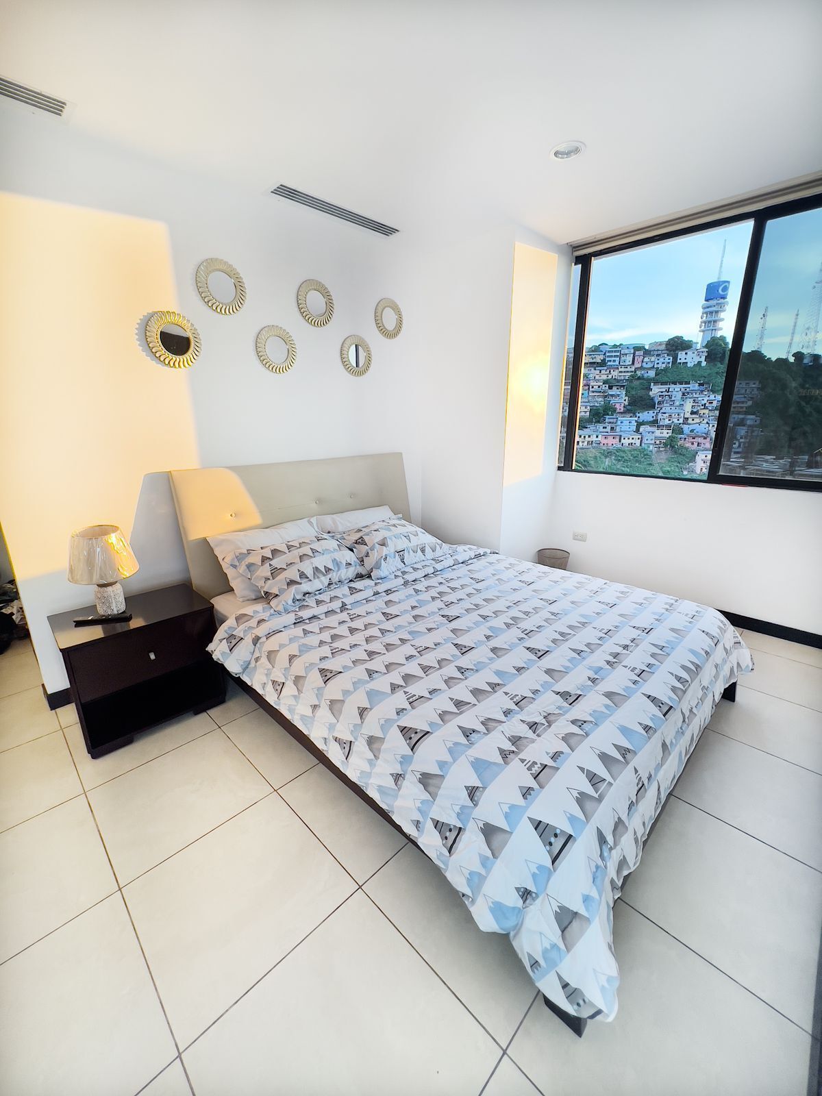 A bedroom with a bed, a nightstand, a lamp, and a large window with a city view.