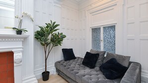 A room with a grey tufted sofa, black cushions, a potted plant, a fireplace, and a window with a mosaic design. - Elegant 4BR Luxury Duplex in chic Upper West Side (New York)