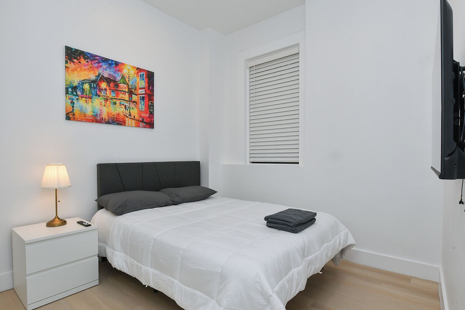 A bedroom with a bed, a bedside table with a lamp, a painting on the wall, a window with blinds, and a TV on the wall.