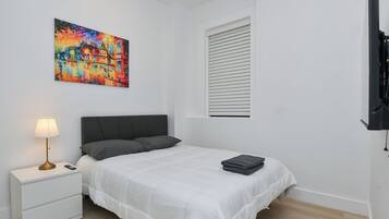 A bedroom with a bed, a bedside table with a lamp, a painting on the wall, a window with blinds, and a TV on the wall.