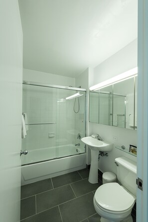 Un baño moderno con ducha envuelta en vidrio, lavamanos pedestal y un inodoro.