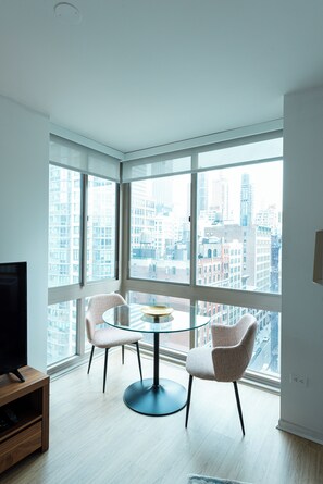 Una habitación de esquina con un ventanal grande, una mesa redonda, dos sillas y un televisor sobre un soporte de madera.