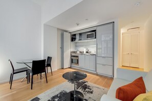 Un espace de vie compact comprenant un coin cuisine, un espace repas avec une table ronde et des chaises, et un canapé avec des coussins.
