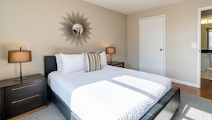 Un dormitorio con una cama grande, dos mesitas de noche con lámparas, un espejo en la pared y una puerta a otra habitación.
