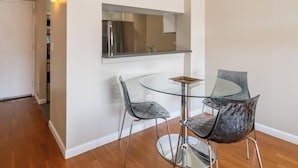 Un comedor con mesa y sillas de vidrio, un refrigerador de acero inoxidable y un estante en la pared.