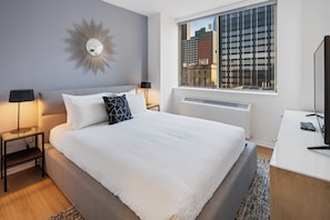 A modern hotel room with a large bed, a desk, a window with a city view, and a decorative wall piece.