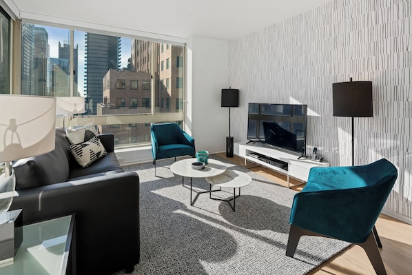 A modern living room with a black sofa, teal armchairs, a white coffee table, and a TV against a patterned wall.