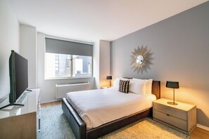 Ein modernes Schlafzimmer mit einem großen Bett, Nachttischen, einem Fernseher und einem Fenster mit Blick auf Gebäude.