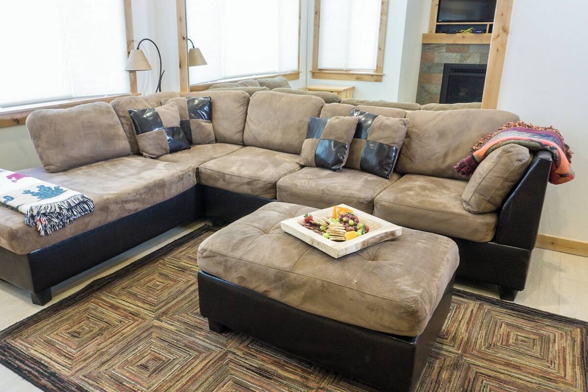 A sectional sofa with a side table and ottoman, a patterned rug, and a small tray of snacks.