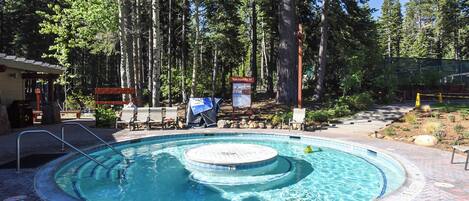 A circular hot tub surrounded by a brick patio, with a wooden deck and lounge chairs in the background.