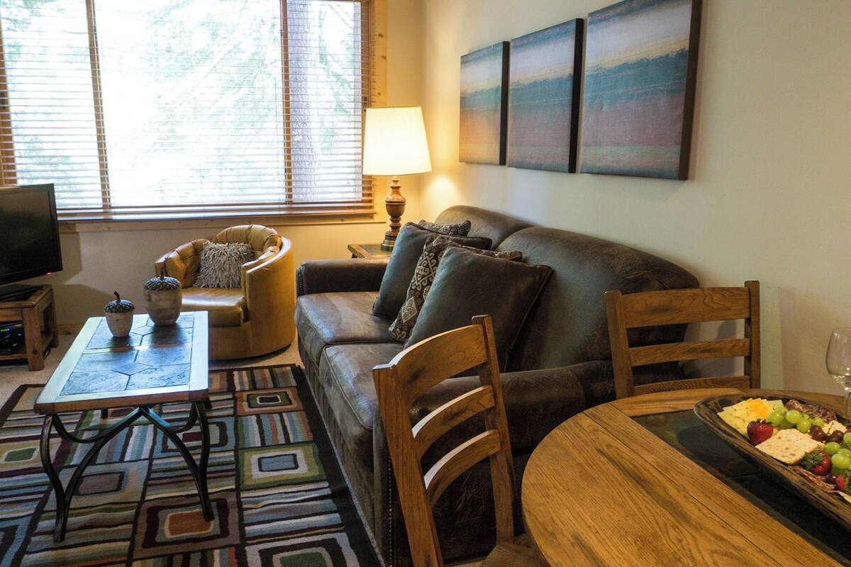 A living room with a leather sofa, a coffee table, a TV on a stand, and a dining table with a fruit platter.