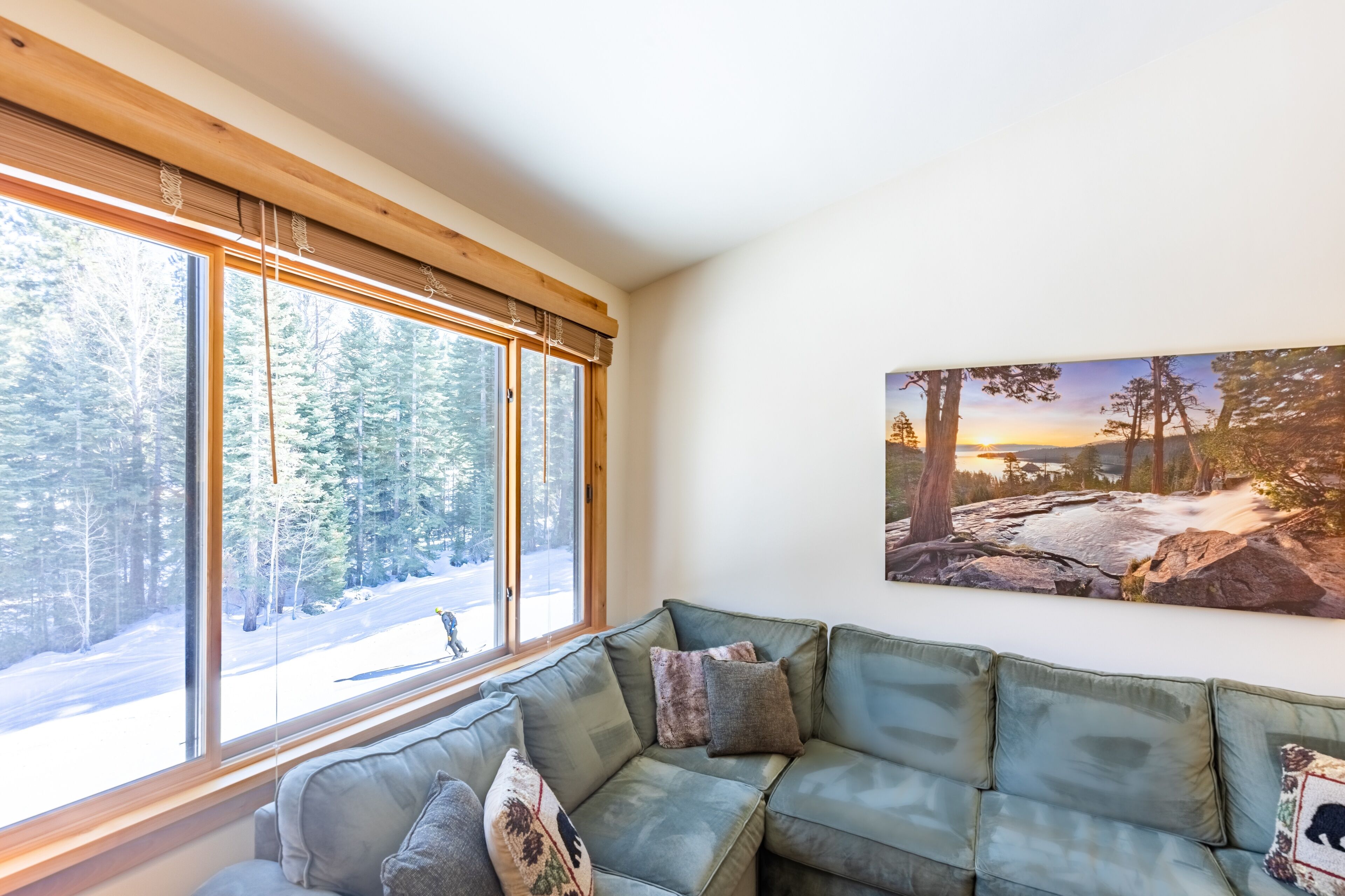 A cozy living room with a green sectional sofa, large windows with a view of a snowy forest, and a painting of a forest scene on the wall.
