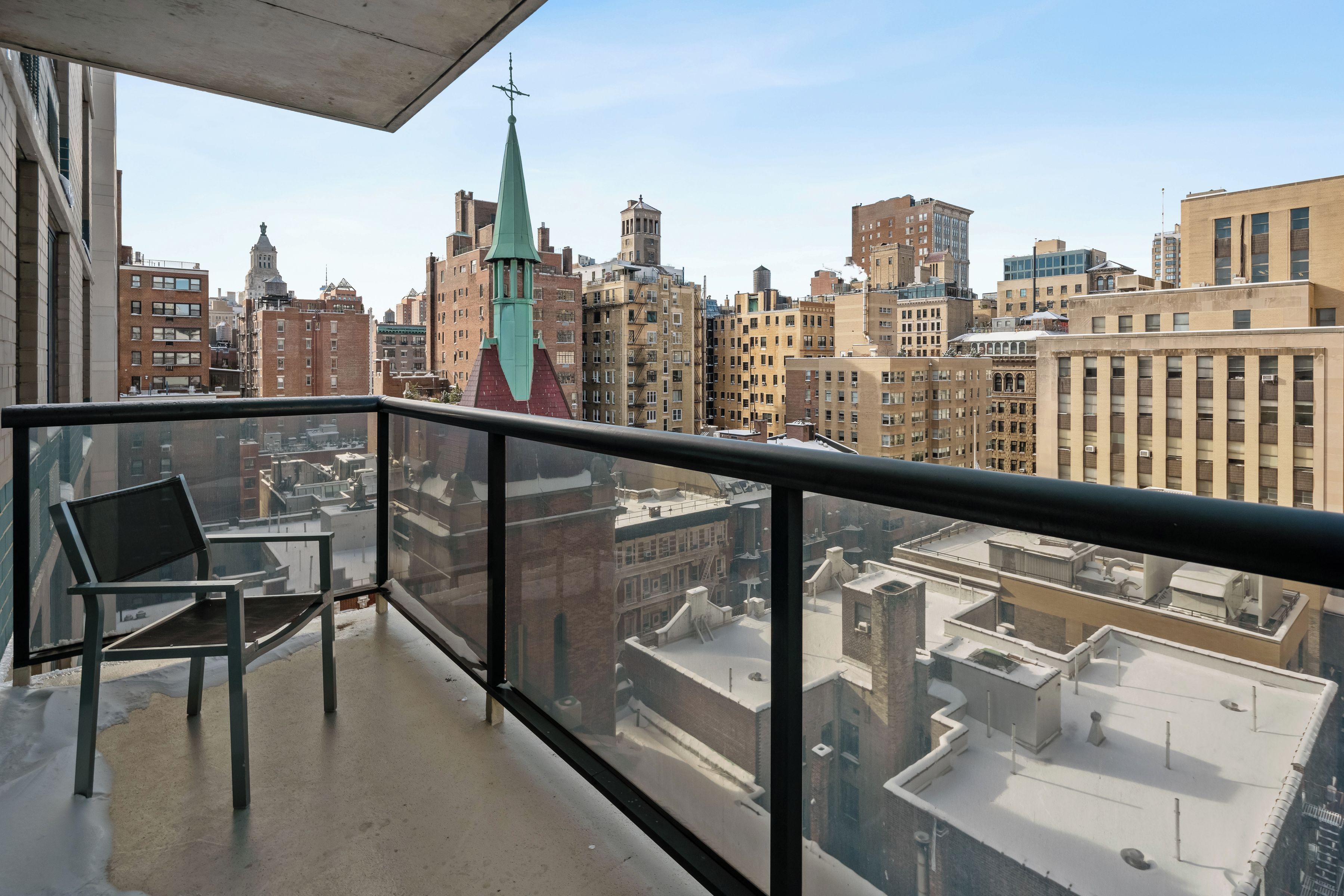 A balcony with a chair, overlooking a cityscape with various buildings and a church steeple.