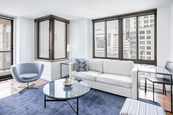 A modern living room with a white sofa, a glass coffee table, a blue armchair, and a large window offering a city view.