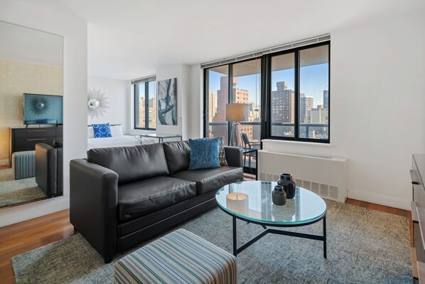 A modern living room with a black leather sofa, a glass coffee table, and a city view through large windows.