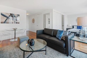 A modern living room with a glass coffee table, a black leather sofa, and a glass dining table. There is a bed with a blue and white patterned pillow in the background.