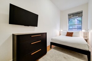 A bedroom with a bed, a dresser, a TV, and a window with a view of buildings.