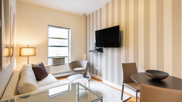 Uma sala de estar moderna com sofá, poltronas, mesa de centro, TV fixada na parede e uma janela que oferece luz natural.