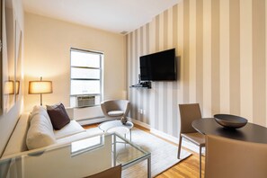 A modern living room with a sofa, armchairs, a coffee table, a TV mounted on the wall, and a window providing natural light.