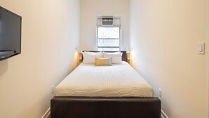 Quarto de hotel estreito com cama, televisão montada e janela com unidade de ar condicionado.