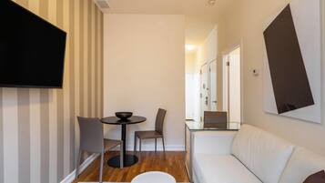 A modern living room with a sofa, a dining table with chairs, a flat-screen TV mounted on the wall, and a glass partition.