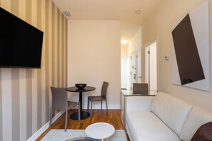 A modern living room with a sofa, a dining table with chairs, a flat-screen TV mounted on the wall, and a glass partition.