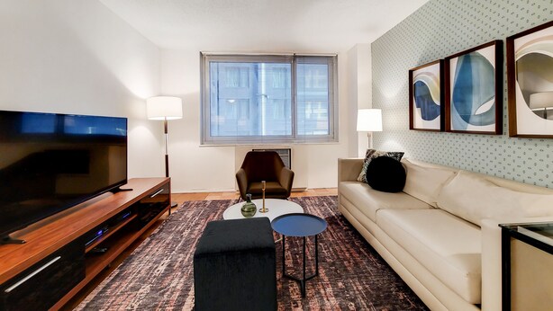 A living room with a TV, sofa, coffee table, and a patterned rug. - Upscale Living | Midtown West |  Doorman & Rooftop (New York)
