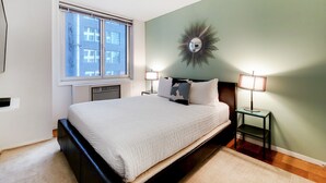 A bedroom with a bed, two bedside tables with lamps, a wall-mounted mirror, and a window with a view of buildings. - Upscale Living | Midtown West |  Doorman & Rooftop (New York)