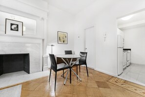 A modern dining area with a marble fireplace, a round table with chairs, and a kitchen with white appliances. - Classic UWS Living | Riverside Park | Pet-Friendly (New York)