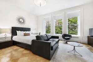 A modern bedroom with a bed, a black sofa, a small round table, and large windows offering a view of greenery outside. - Classic UWS Living | Riverside Park | Pet-Friendly (New York)