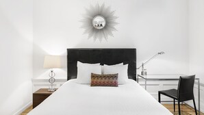 Un letto rifatto con lenzuola bianche, uno specchio decorativo a parete, una lampada e una sedia.