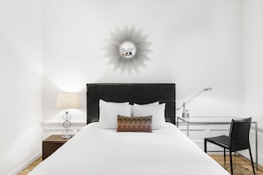 A neatly made bed with white bedding, a decorative wall mirror, a lamp, and a chair.