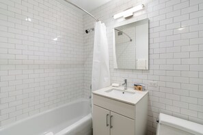 A modern bathroom with white subway tiles, a white bathtub, a vanity with a sink, a mirror, and a shower curtain. - Stunning Greenwich Village Studio with Amenities (New York)