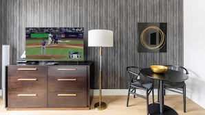 Un saló modern amb televisió de pantalla plana que mostra un partit de beisbol, una làmpada de peu, una taula de dinar amb un bol i un armari de fusta fosca amb calaixos.