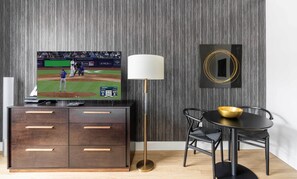 A modern living room with a flat-screen TV displaying a baseball game, a floor lamp, a dining table with a bowl, and a dark wooden cabinet with drawers.