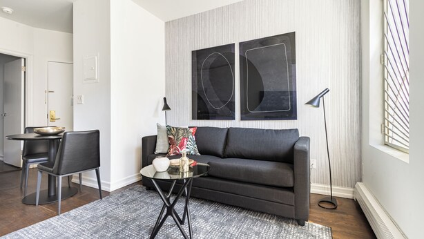 Een moderne woonkamer met een donkergrijze bank, een glazen salontafel en een vloerkleed met dessin. Er is een zithoek met een kleine tafel en stoelen, en een staande lamp.