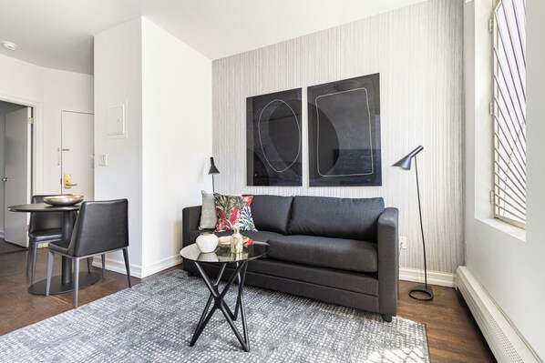 A modern living room with a dark grey sofa, a glass coffee table, and a patterned rug. There is a dining area with a small table and chairs, and a floor lamp.