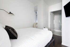 A modern bedroom with a large bed, a bedside table, a lamp, and a shower area visible through a glass door.