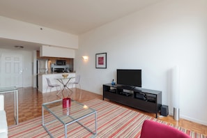 A modern living room with a glass coffee table, a red chair, a TV on a dark stand, and a kitchen in the background.