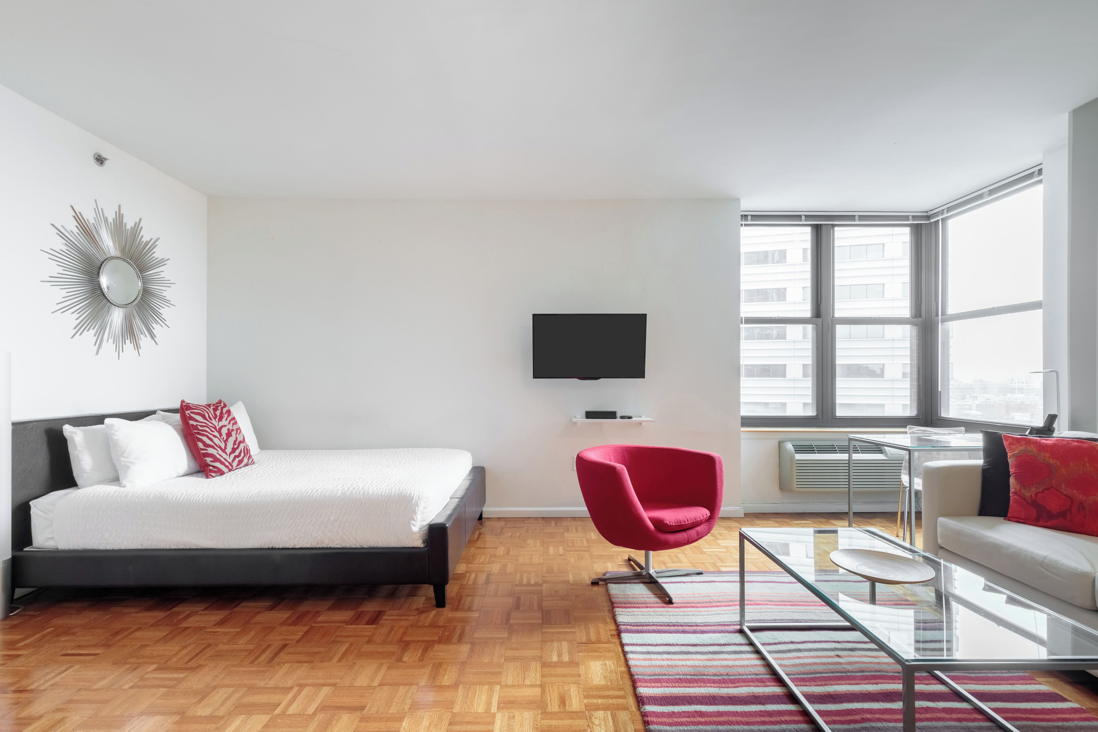 Een moderne hotelkamer met een bed, een rode stoel, een glazen salontafel, een bank, een televisie en grote ramen.