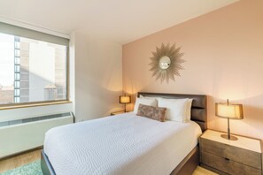 Una cama bien tendida con edredón blanco, dos lámparas de noche, un espejo decorativo y una ventana con vistas a edificios.