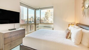 A modern bedroom with a large bed, a TV on a wooden dresser, and a city view through large windows. - Centrally Located Chelsea Luxe One Bedroom (New York)
