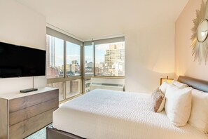 A modern bedroom with a large bed, a TV on a wooden dresser, and a city view through large windows.