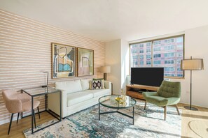 A modern living room with a white sofa, a glass coffee table, a green armchair, a TV, and artwork on the wall.