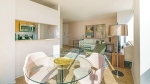 A modern living room with a glass table, two chairs, a sofa, and a coffee table. There is a kitchen area with a microwave and cabinets. The room has a wooden floor and a large window providing natural light. - Centrally Located Chelsea Luxe One Bedroom (New York)
