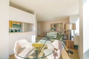 A modern living room with a glass table, two chairs, a sofa, and a coffee table. There is a kitchen area with a microwave and cabinets. The room has a wooden floor and a large window providing natural light.