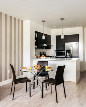 Una cocina moderna con una isla central, armarios negros, electrodomésticos de acero inoxidable y un comedor con mesa de vidrio y sillas negras.