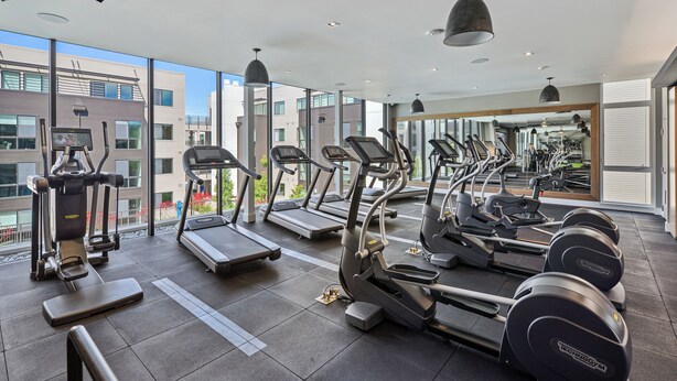 Un gimnasio moderno con cintas de correr, elípticas y máquinas de pesas, amplias ventanas y vistas de la ciudad.